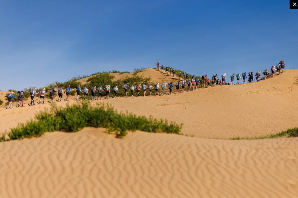 Long cordon de concurrents de la Gobi March, sac au dos, gravissant une dune de sable sous un ciel bleu lors de l'étape longue